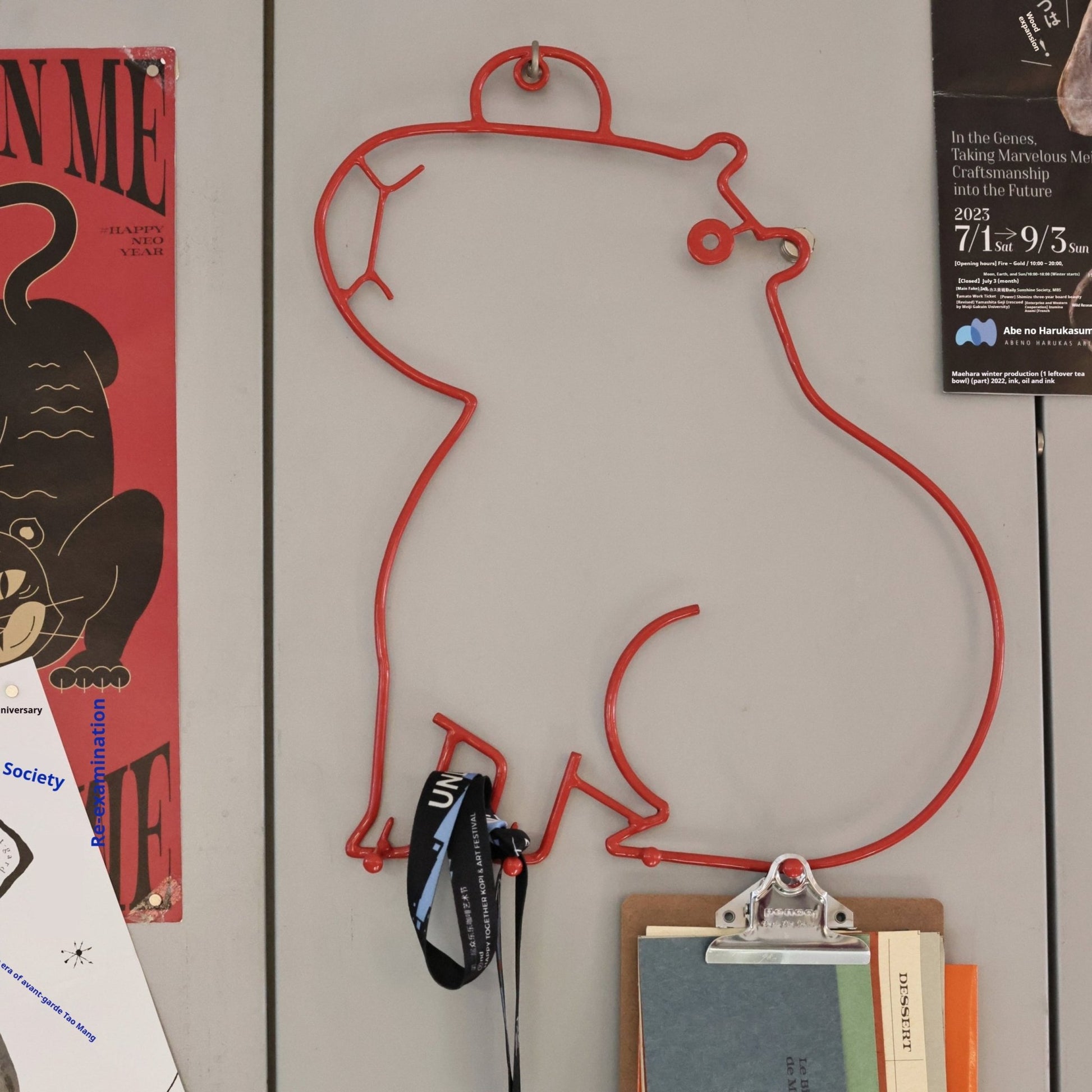 Red wire wall hook shaped like a person with items attached on a gray wall.
