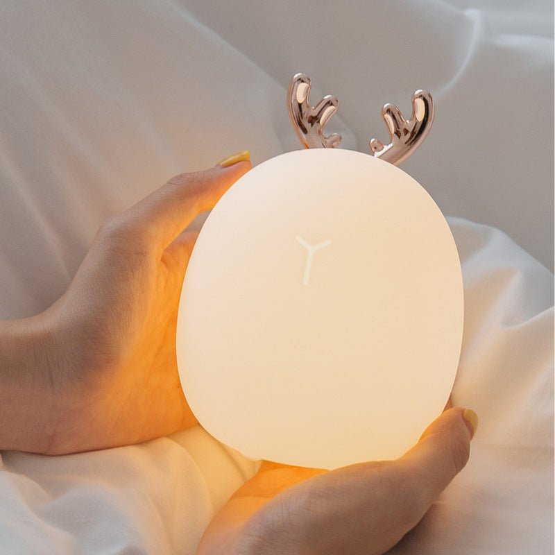 Hand holding a round, glowing night light with deer antlers on a soft fabric background