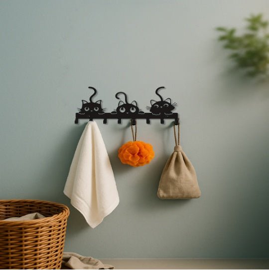 Black cat-shaped hooks on a wall with a towel, orange item, and beige bag hanging from them.