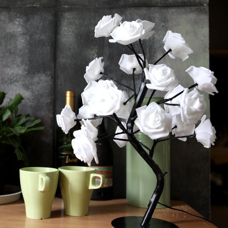 Decorative white rose tree on a table with green mugs and a bottle in the background.