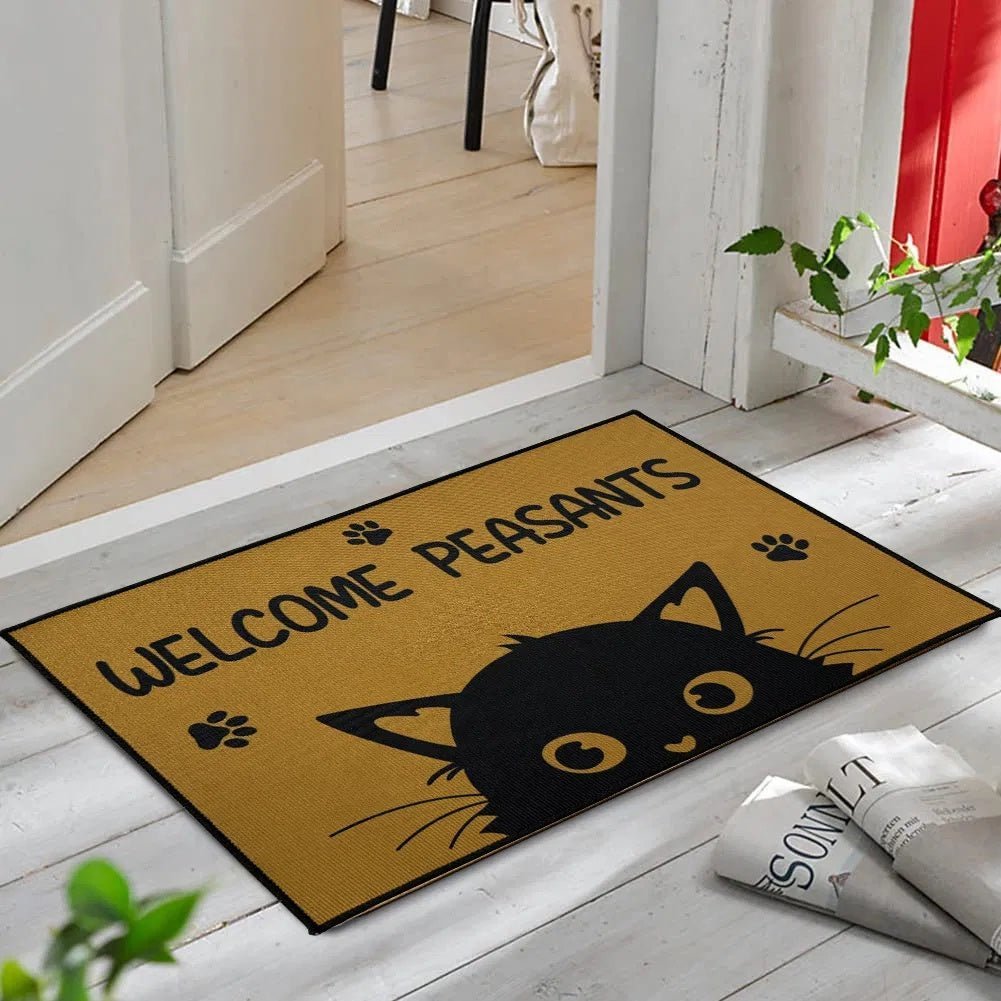 Doormat with a black cat design and text 'Welcome Peasants' on a wooden floor.